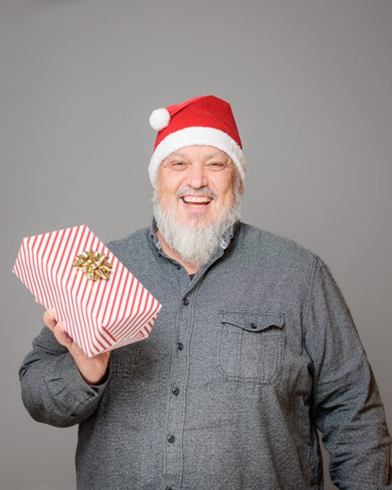 Älterer Mann mit weißem Bart, trägt Weihnachtsmütze, lächelt und hält ein rot-weiß verpacktes Geschenk mit goldener Schleife.