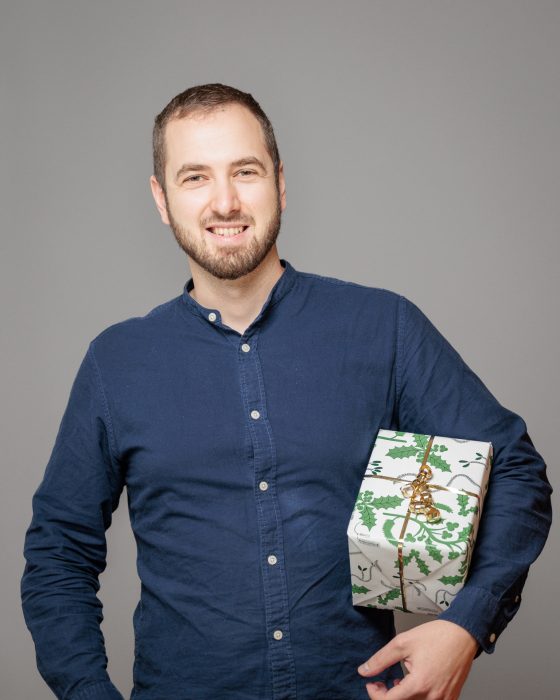 Mann in dunkelblauem Hemd hält lächelnd ein Geschenk mit grün-weißem Papier und goldenem Band auf grauem Hintergrund.