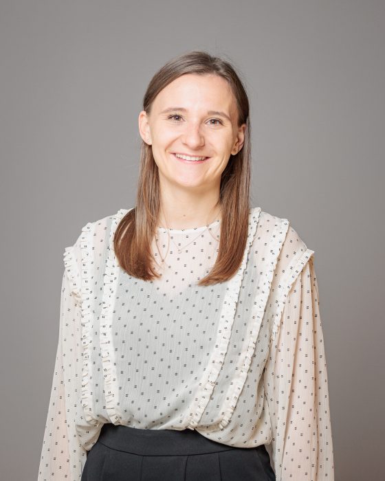 Eine lächelnde Frau mit langen, glatten Haaren trägt eine gepunktete Bluse mit Rüschen vor einem grauen Hintergrund.