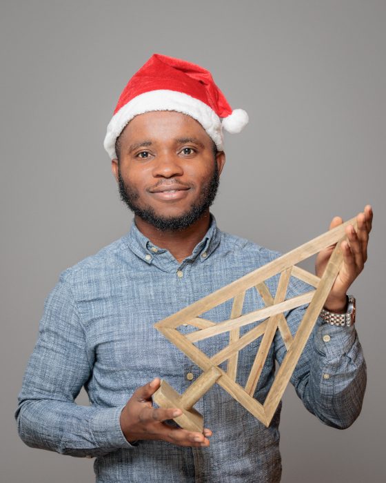 Ein lächelnder Mann trägt eine rote Weihnachtsmütze und hält eine dekorative, hölzerne Dreiecksform, die an einen Weihnachtsbaum erinnert.