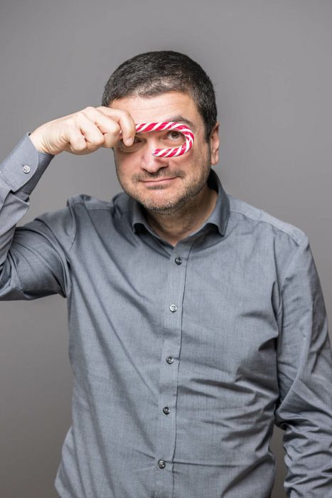 Ein Mann in einem grauen Hemd hält einen rot-weiß gestreiften Zuckerstab vors Auge und lächelt in die Kamera.