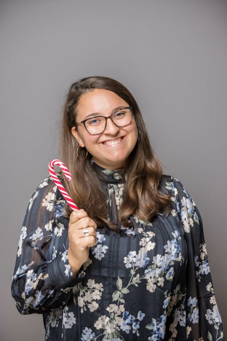 Eine lächelnde Frau mit Brille und langem braunem Haar hält einen rot-weißen Zuckerstab. Sie trägt eine Blumenbluse.