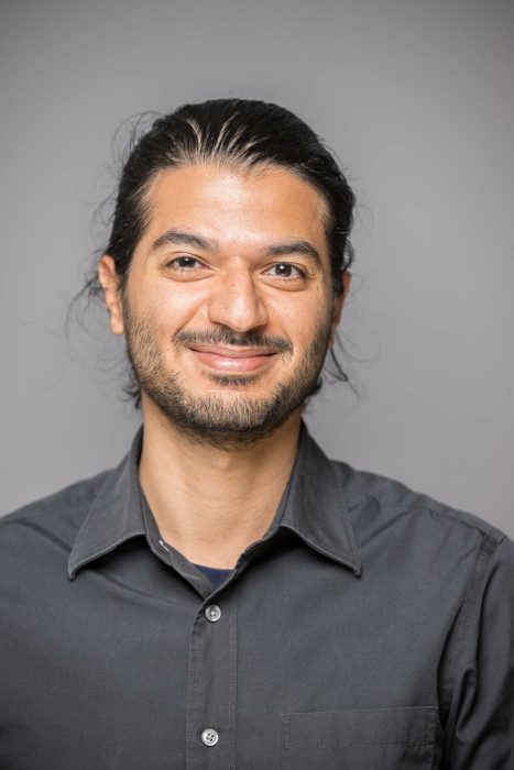 Ein Mann mit dunklen Haaren, die nach hinten gebunden sind, trägt ein dunkles Hemd und lächelt in die Kamera vor einem grauen Hintergrund.