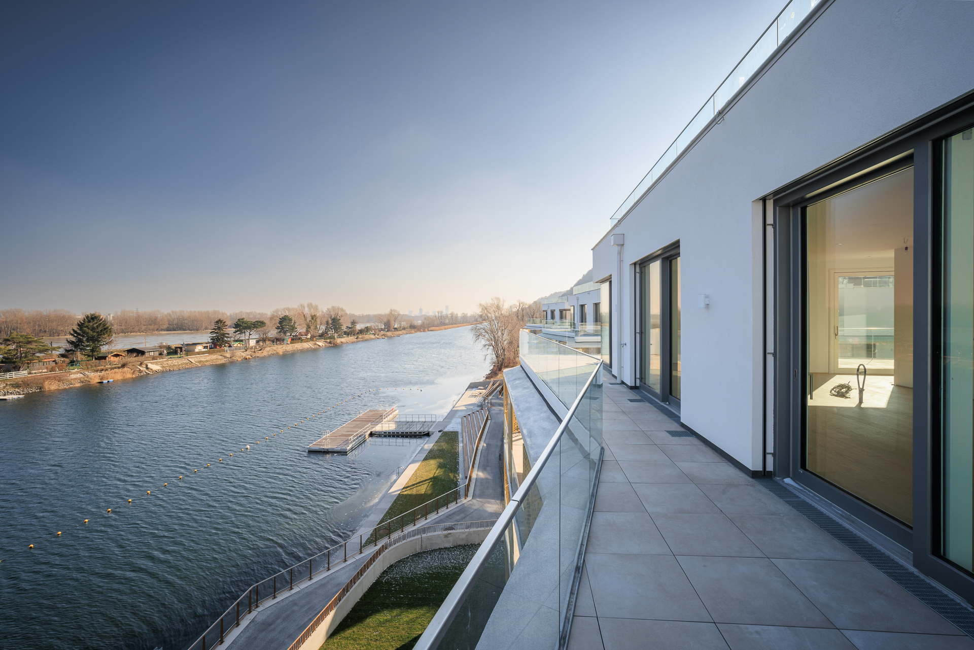 Ein moderner Balkon mit Glasgeländer verläuft entlang eines weißen Gebäudes, mit Blick auf einen breiten Fluss und eine mit Bäumen gesäumte Uferlandschaft unter einem klaren blauen Himmel.