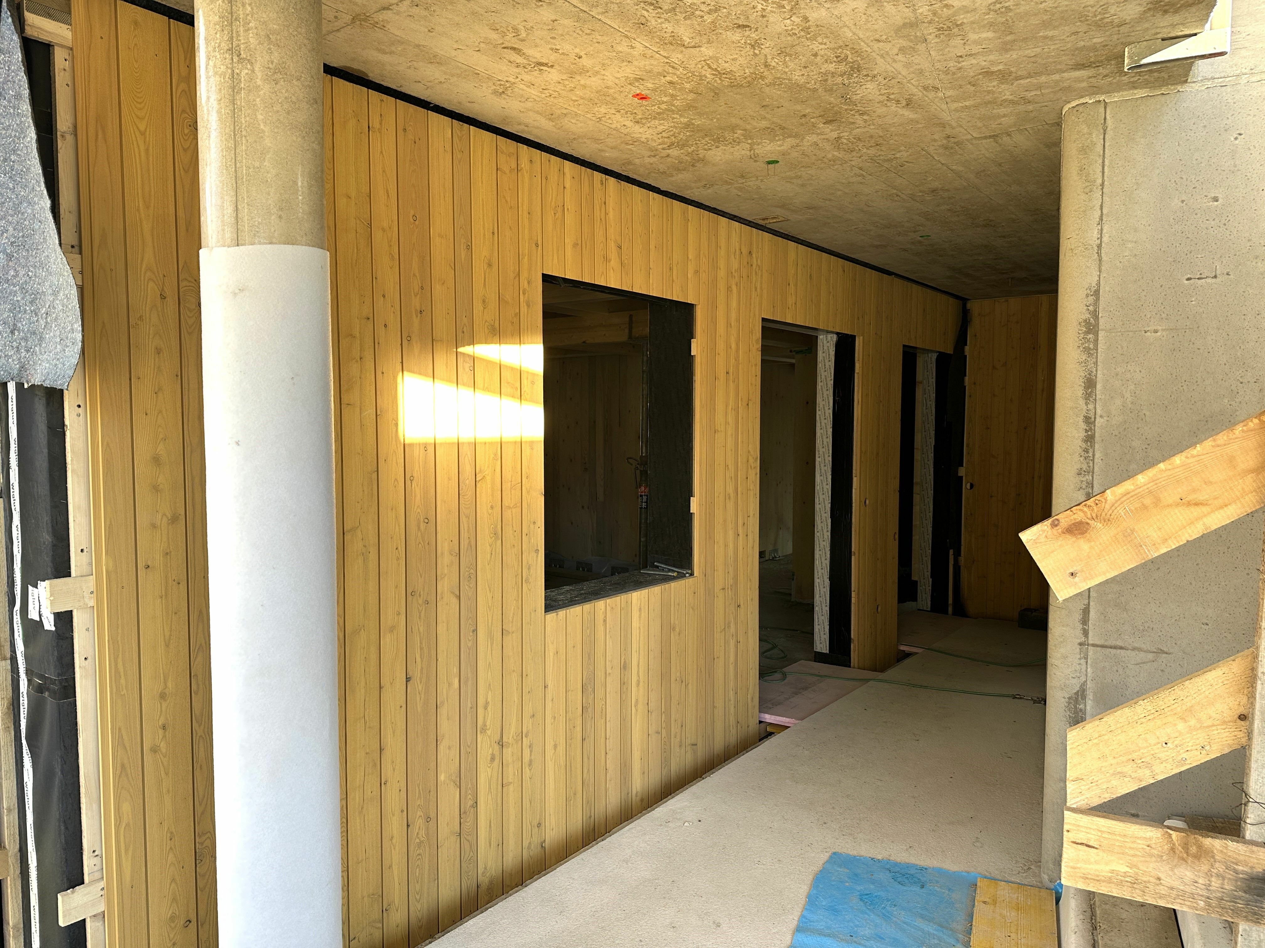 Ein unfertiger Innenkorridor mit Holzwandverkleidung, Betonwänden und offenen Türrahmen. Der Boden ist teilweise mit Schutzmaterial bedeckt.