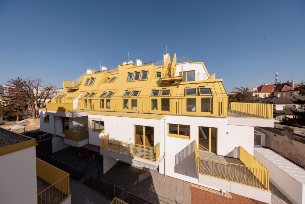 Das Bild zeigt ein modernes, mehrstöckiges Wohngebäude mit gelben Akzenten, großen Fenstern und Balkonen, umgeben von einem blauen Himmel und benachbarten Gebäuden.