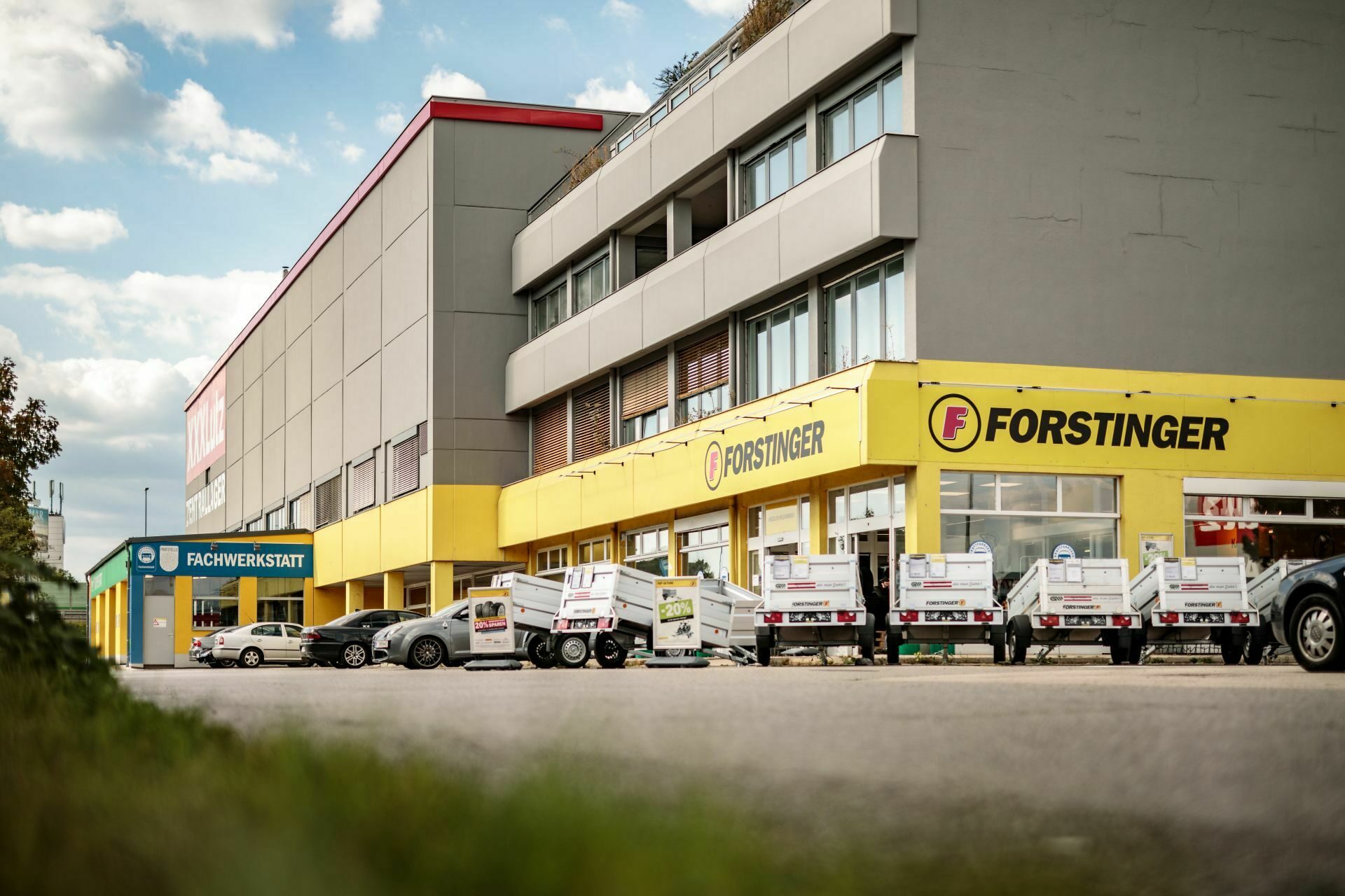 Außenansicht eines Gebäudes mit gelbem Forstinger-Schild, mehreren Anhängern vor dem Eingang und einem Schild mit der Aufschrift "Fachwerkstatt".