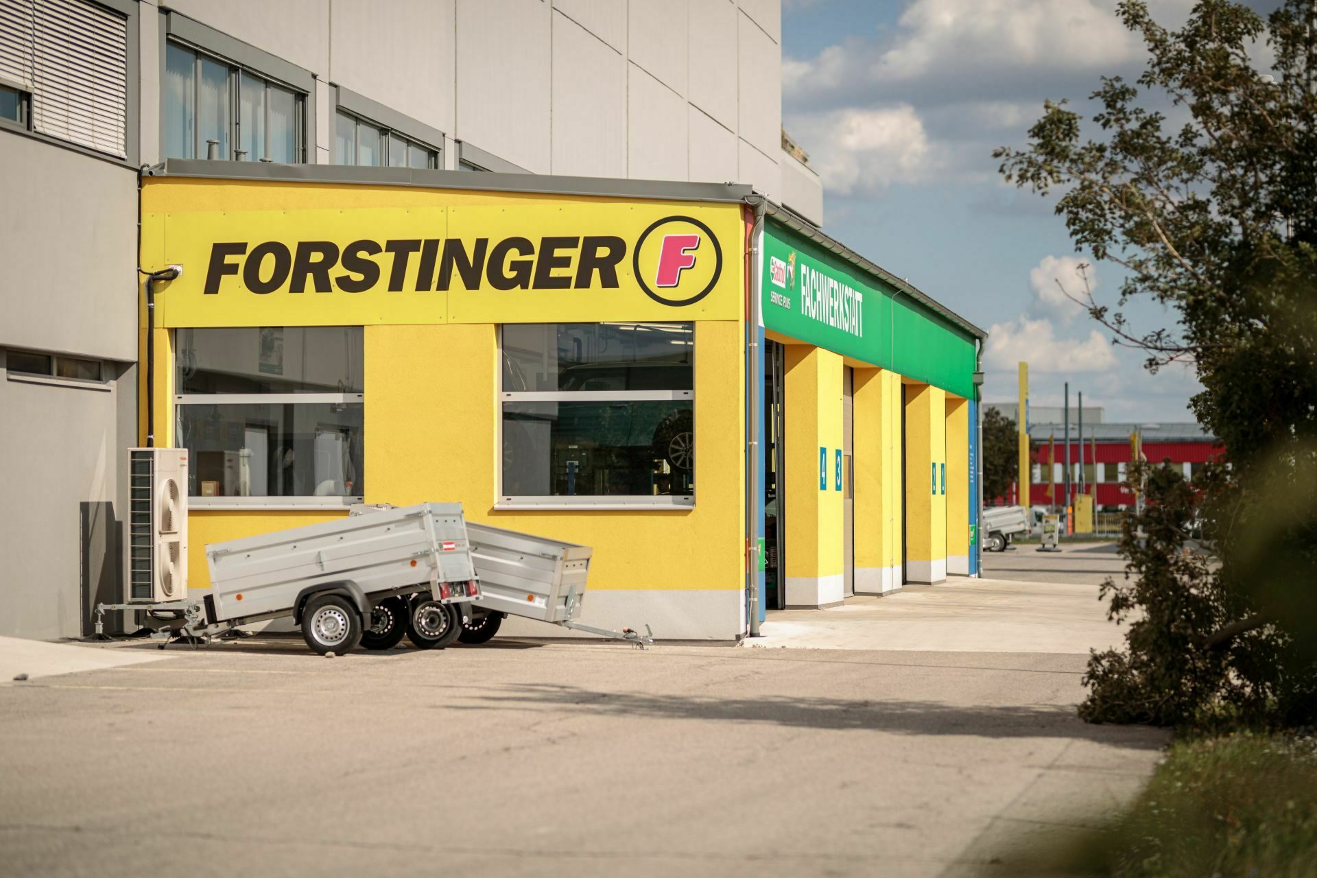 Gelbes Gebäude mit der Aufschrift "FORSTINGER", einem Anhänger davor und einer Werkstatt mit grünen Details auf der rechten Seite.