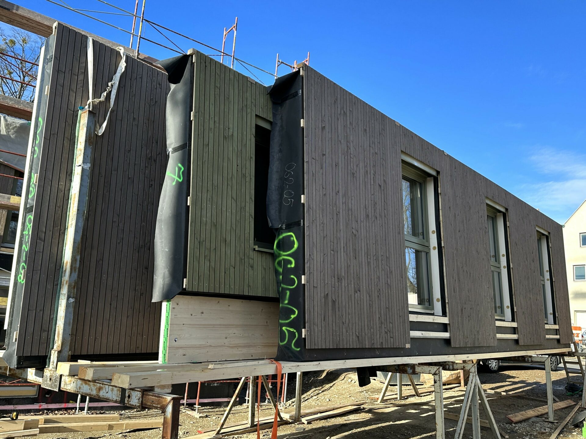 Ein vorgefertigtes Gebäudemodul mit Holzverkleidung und mehreren Fenstern steht auf Metallstützen auf einer Baustelle unter blauem Himmel.